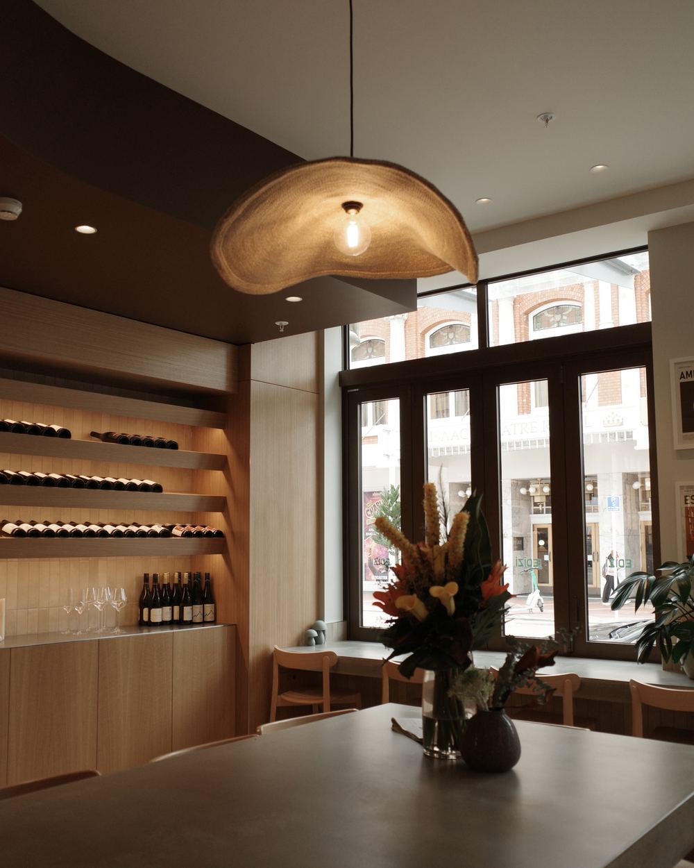 A Christchurch wine room: lit oak shelves of bottles, a woven pendant light and a flower arrangement on the table